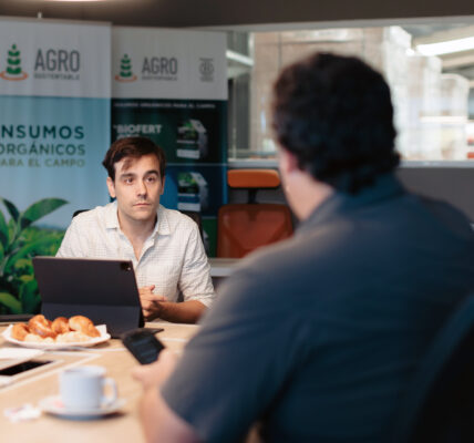 Matías Imperiale en una reunión de trabajo en la sede de Agro Sustentable, frente a una notebook, con banners institucionales de insumos orgánicos al fondo.