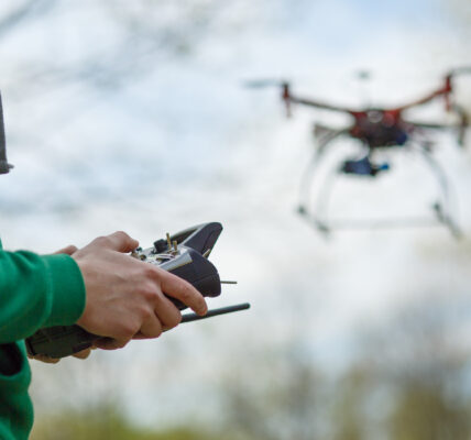 Persona con buzo verde controla un dron agrícola en vuelo mediante un control remoto, en un entorno natural al aire libre.