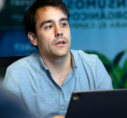 Hombre concentrado mirando una tableta digital en un ambiente de oficina con carteles informativos de fondo.