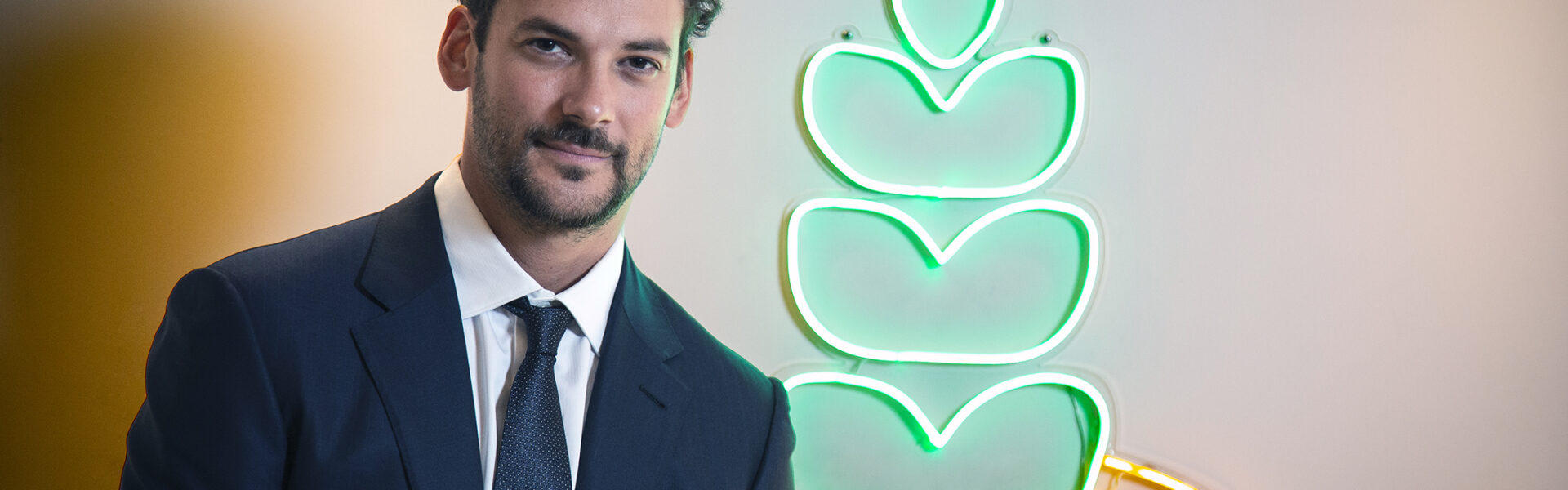 Joaquín Basanta con traje oscuro posando frente a un ícono luminoso verde y amarillo en forma de planta estilizada sobre fondo claro.