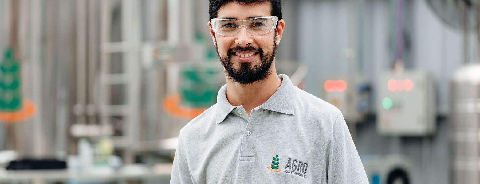 Técnico de Agro Sustentable con gafas de protección en planta de producción de bioinsumos, sonriendo frente a tanques industriales.
