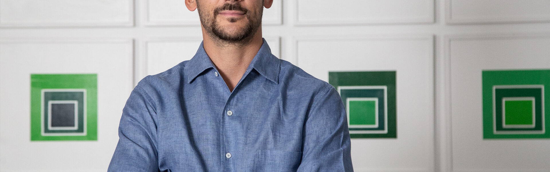 Retrato de un hombre joven de camisa azul, de pie con los brazos cruzados frente a una pared con obras geométricas en tonos verdes.