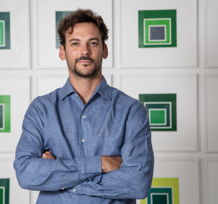 Retrato de un hombre joven de camisa azul, de pie con los brazos cruzados frente a una pared con obras geométricas en tonos verdes.