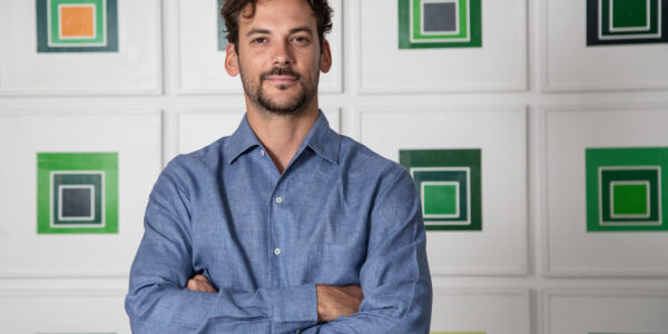 Retrato de un hombre joven de camisa azul, de pie con los brazos cruzados frente a una pared con obras geométricas en tonos verdes.