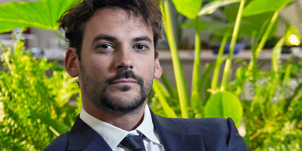 Hombre joven en traje formal, posando frente a un fondo de plantas verdes en un entorno profesional.