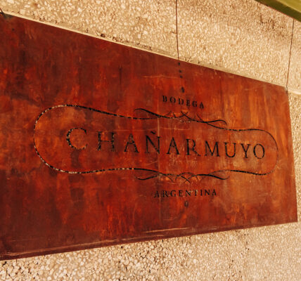 Cartel de chapa oxidada con el nombre “Bodega Chañarmuyo” colgado sobre una pared texturada.