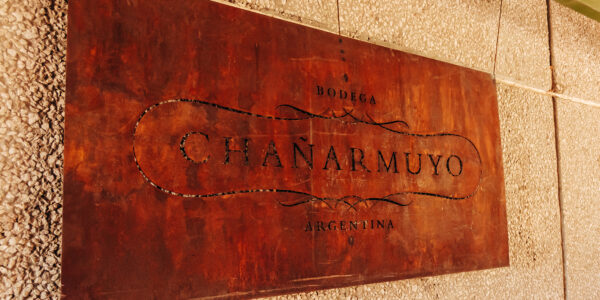 Cartel de chapa oxidada con el nombre “Bodega Chañarmuyo” colgado sobre una pared texturada.