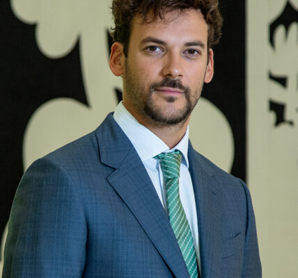 Joaquín Basanta con traje azul y corbata verde, posando frente a una obra gráfica en blanco y negro.