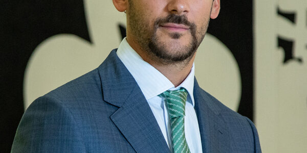 Joaquín Basanta con traje azul y corbata verde, posando frente a una obra gráfica en blanco y negro.