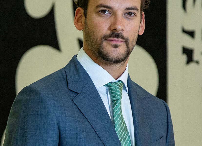 Joaquín Basanta con traje azul y corbata verde, posando frente a una obra gráfica en blanco y negro.