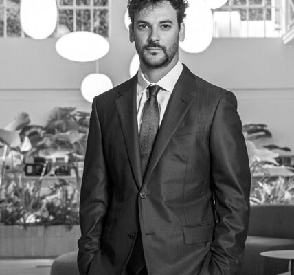 Hombre de traje oscuro en un entorno moderno con vegetación interior y lámparas redondas, mirando a cámara con gesto serio.