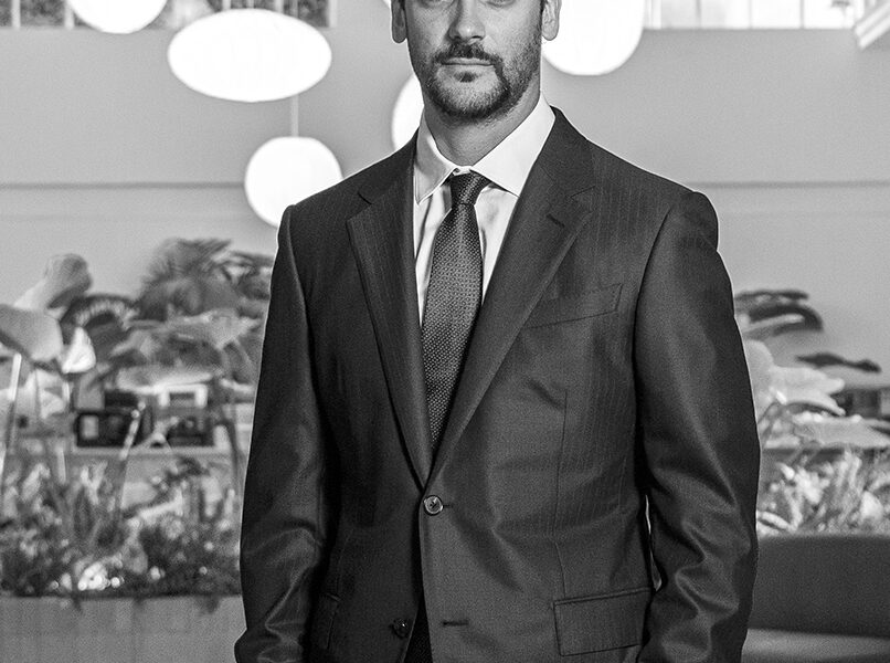 Hombre de traje oscuro en un entorno moderno con vegetación interior y lámparas redondas, mirando a cámara con gesto serio.