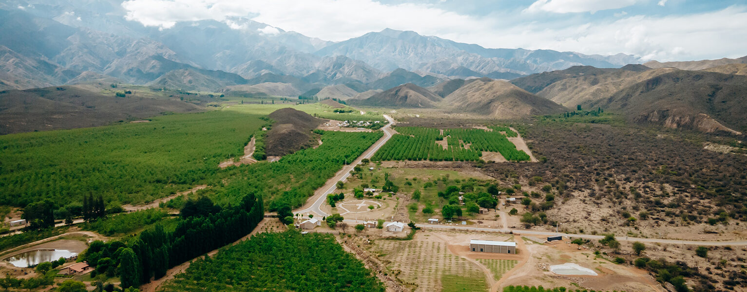 Vista aérea de un valle agrícola rodeado de montañas con cultivos verdes, caminos rurales y cielo parcialmente nublado.
