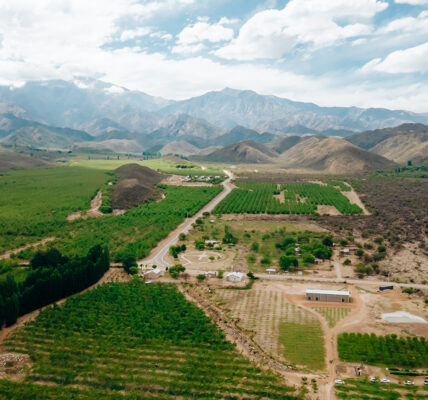 Vista aérea de un valle agrícola rodeado de montañas con cultivos verdes, caminos rurales y cielo parcialmente nublado.