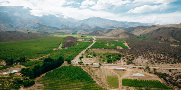 Vista aérea de un valle agrícola rodeado de montañas con cultivos verdes, caminos rurales y cielo parcialmente nublado.
