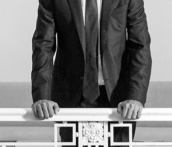 Hombre de traje oscuro apoyado sobre una baranda blanca, en un espacio interior iluminado, mirando al frente con actitud segura.