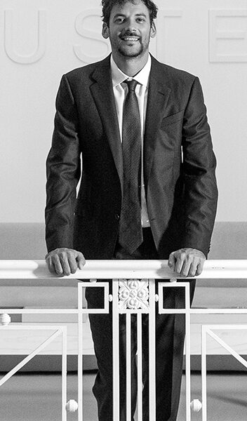 Hombre de traje oscuro apoyado sobre una baranda blanca, en un espacio interior iluminado, mirando al frente con actitud segura.